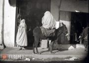 Two figures in burnouses, with a donkey in front of a shop cafe